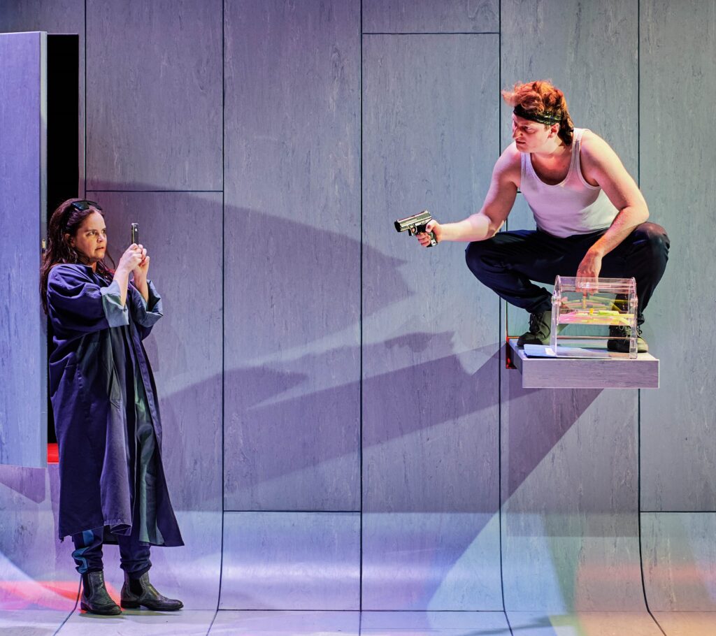 Szene aus dem Theaterstück „Jeeps“ in der Inszenierung durch die Württembergische Landesbühne Esslingen: Vor einer grauen Wand filmt eine Figur mit dem Handy eine andere Figur, die mit gezückter Pistole auf einem Tresen hockt. (Bild: Patrick Pfeiffer)