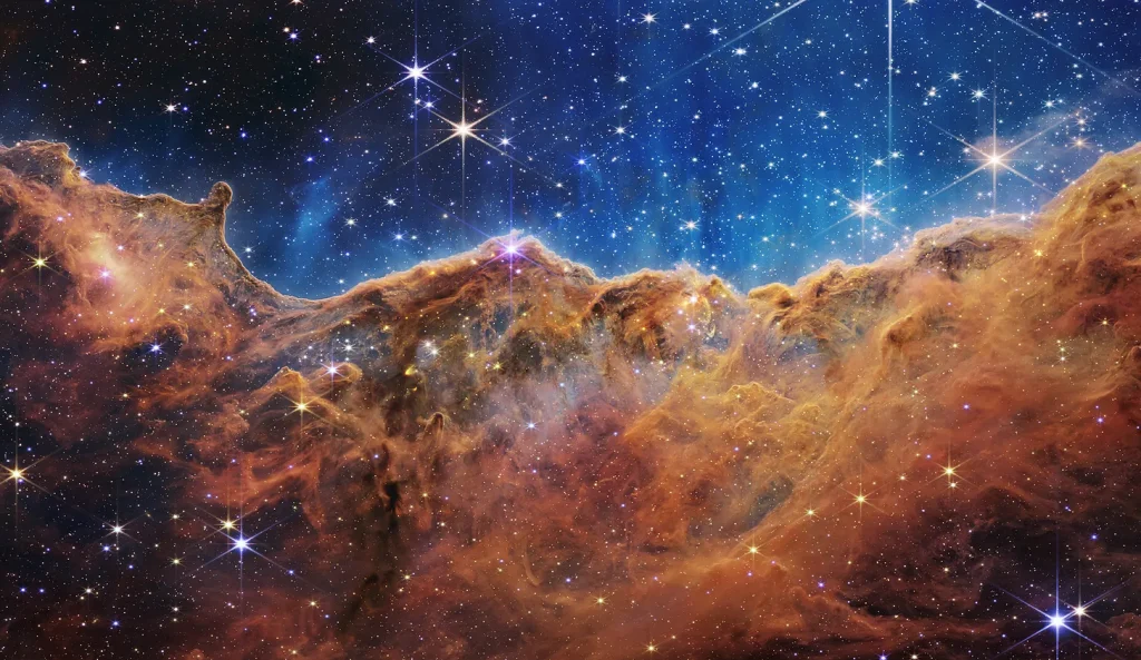 Carina-Nebel im Carina-Sternbild, aufgenommen vom James-Webb-Teleskop: eine hellbraun-goldene, kosmische "Nebelwolke", die etwa 3/4 des Bildes ausmacht. Darin und darüber sind Sterne und das teils dunkelbläuliche Schwarz des Weltalls zu sehen.