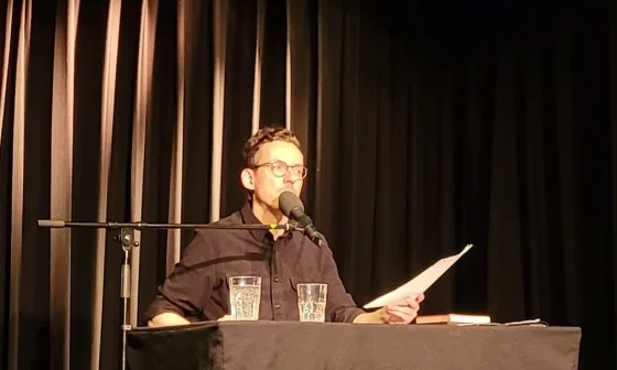 Auf einer dunklen Bühne sitzt Paul Bokowsi an einem schwarzen Tisch. Er trägt ein schwarzes Hemd, hält ein Blatt Papier in der Hand, schaut zur Seite und spricht ins Mikrofon.