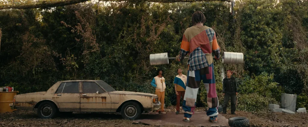 Szene aus der Prime-Video-Serie I'm A Virgo: Die vier Meter große Hauptfigur steht mit einer auf sie zugeschnittenen Stangen-Hantel in einem Garten mit sehr hoher Hecke im Hintergrund, man sieht die Figur von hinten. Links neben der Figur steht ein heruntergekommenes Auto. Vor der Figur stehen drei Menschen, die zu ihr hochschauen.