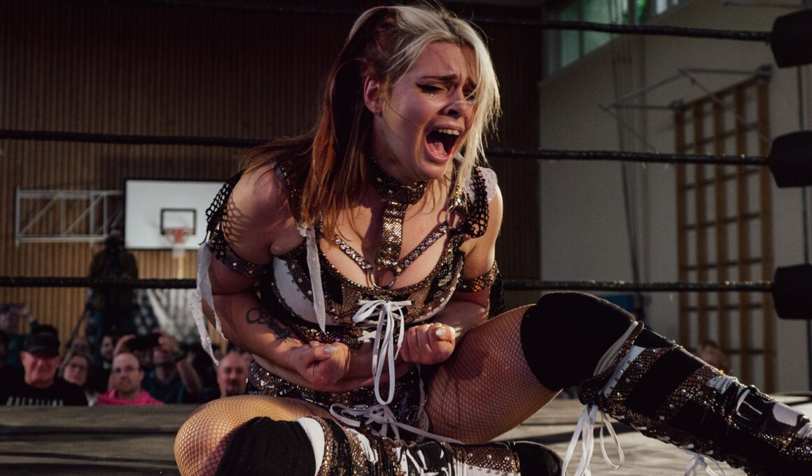 Die Wrestlerin Calypso sitzt bei der Frauen-Wrestling-Veranstaltung „Breaking Barriers“ auf dem Ringboden und ballt schreiend die Fäuste. Sie trägt schwarz-weiß-goldene Wrestling-Kleidung. (Foto: Nadine Veigel/Photos N Effect)