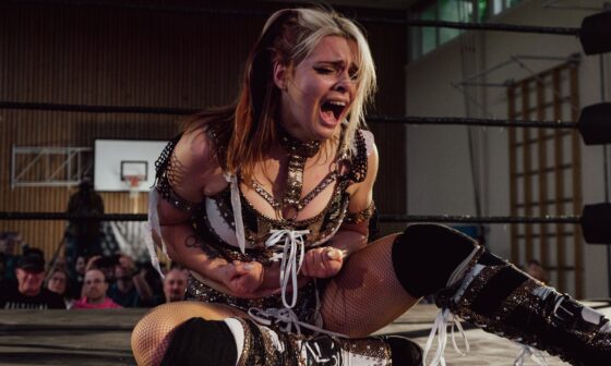 Die Wrestlerin Calypso sitzt bei der Frauen-Wrestling-Veranstaltung „Breaking Barriers“ auf dem Ringboden und ballt schreiend die Fäuste. Sie trägt schwarz-weiß-goldene Wrestling-Kleidung. (Foto: Nadine Veigel/Photos N Effect)