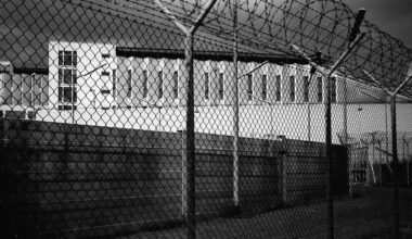 Schwarzweiß-Foto der Justizvollzugsanstalt Stuttgart-Stammheim, fotografiert von außen direkt vorm Stacheldraht-Zaun. Zu sehen sind die Mauer und ein Gefängnistrakt.
