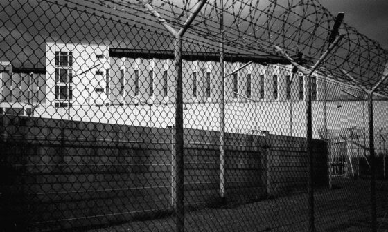 Schwarzweiß-Foto der Justizvollzugsanstalt Stuttgart-Stammheim, fotografiert von außen direkt vorm Stacheldraht-Zaun. Zu sehen sind die Mauer und ein Gefängnistrakt.
