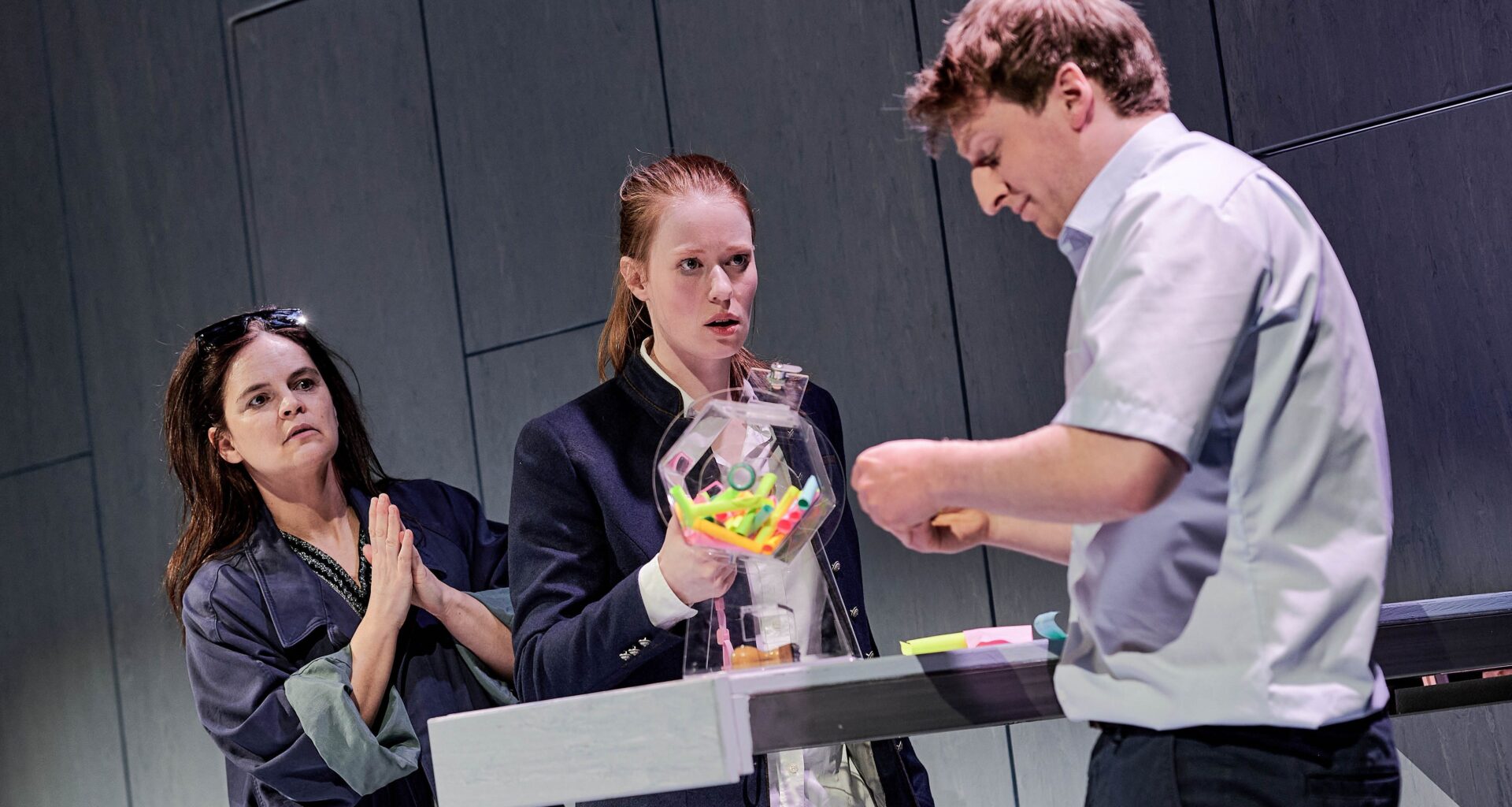 Szene aus dem Theaterstück „Jeeps“ in der Inszenierung durch die Württembergische Landesbühne Esslingen: Vor einer grauen Wand stehen drei Personen vor und hinter einem Amtsschalter, auf dem sich eine Lostrommel befindet. (Bild: Patrick Pfeiffer)