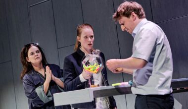 Szene aus dem Theaterstück „Jeeps“ in der Inszenierung durch die Württembergische Landesbühne Esslingen: Vor einer grauen Wand stehen drei Personen vor und hinter einem Amtsschalter, auf dem sich eine Lostrommel befindet. (Bild: Patrick Pfeiffer)