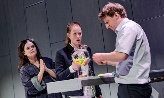 Szene aus dem Theaterstück „Jeeps“ in der Inszenierung durch die Württembergische Landesbühne Esslingen: Vor einer grauen Wand stehen drei Personen vor und hinter einem Amtsschalter, auf dem sich eine Lostrommel befindet. (Bild: Patrick Pfeiffer)