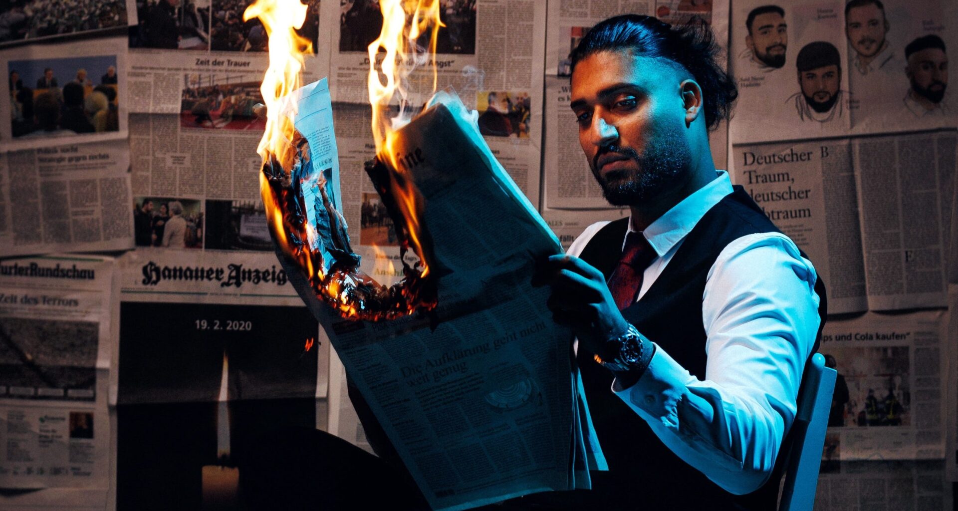 Said Etris Hashemi liest sitzend eine brennende Zeitung. Im Hintergrund hängen Zeitungsartikel über den rechtsterroristischen Anschlag in Hanau. (Foto: Ondro)
