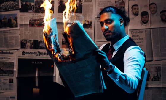 Said Etris Hashemi liest sitzend eine brennende Zeitung. Im Hintergrund hängen Zeitungsartikel über den rechtsterroristischen Anschlag in Hanau. (Foto: Ondro)