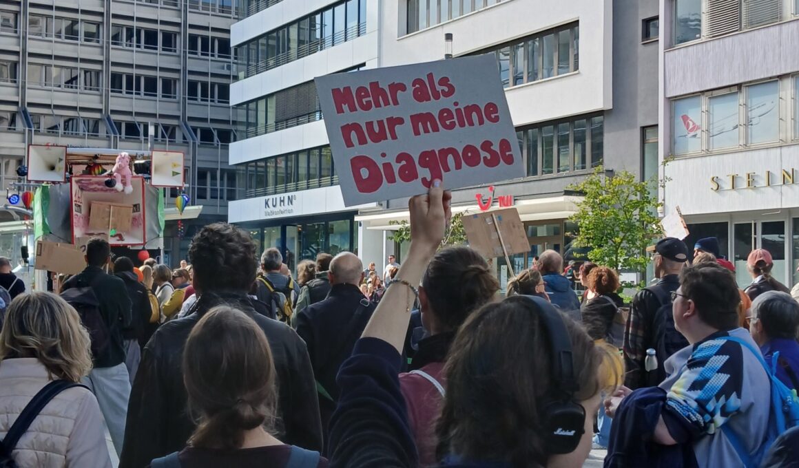 Mad-Pride-Demo in Stuttgart 2025. Jemand hält eine Schild hoch, auf dem in roter Schrift steht: Mehr als meine Diagnose.