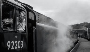 Schwarz-Weiß-Foto einer Dampflok von schräg vorne an einem Bahnhof. Ein Lokführer schaut aus dem Fenster nach vorne. (Foto: Lee Hull auf Unsplash)