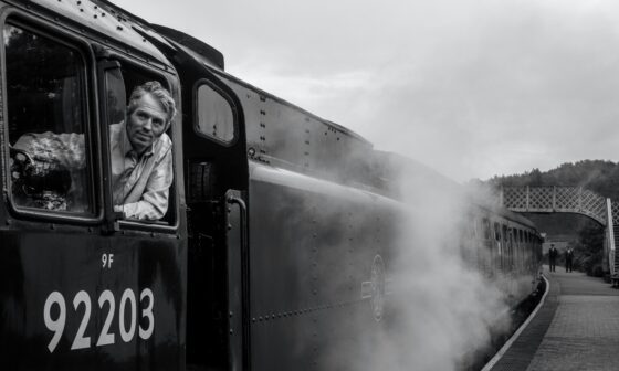 Schwarz-Weiß-Foto einer Dampflok von schräg vorne an einem Bahnhof. Ein Lokführer schaut aus dem Fenster nach vorne. (Foto: Lee Hull auf Unsplash)