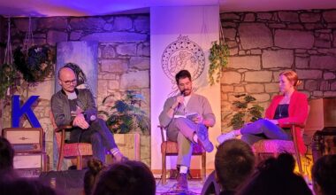 Die Autoren Daniel Stähr und Simon Sahner sitzen mit der Moderatorin Martha Routen auf der Bühne des Freiburger Kulturaggregats. Sahner liest aus dem Sachbuch „Die Sprache des Kapitalismus“ vor. (Foto: vliestext)