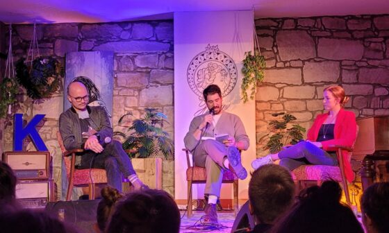 Die Autoren Daniel Stähr und Simon Sahner sitzen mit der Moderatorin Martha Routen auf der Bühne des Freiburger Kulturaggregats. Sahner liest aus dem Sachbuch „Die Sprache des Kapitalismus“ vor. (Foto: vliestext)