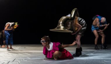 Vor schwarzem Hintergrund und auf silbernem Boden sind vier SHELTER-Tänzer*innen zu sehen. Sie sind bunt gekleidet. Eine*r sitzt in Flughaltung auf dem Boden. Über ihr*ihm hat sich die Tänzer*in mit Sousafon gebeugt. (Foto: Fernanda Tafner)