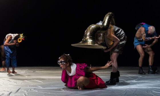 Vor schwarzem Hintergrund und auf silbernem Boden sind vier SHELTER-Tänzer*innen zu sehen. Sie sind bunt gekleidet. Eine*r sitzt in Flughaltung auf dem Boden. Über ihr*ihm hat sich die Tänzer*in mit Sousafon gebeugt. (Foto: Fernanda Tafner)