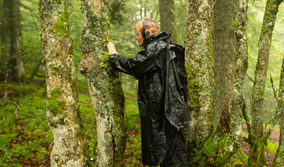 In schwarzer Regenkleidung steht die Komponistin Kirsten Reese in einer Baumgruppe und berührt die Rinde eines Baumes. Sie trägt Kopfhörer. (Foto: Marc Folcher)