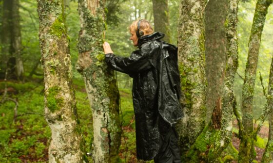 In schwarzer Regenkleidung steht die Komponistin Kirsten Reese in einer Baumgruppe und berührt die Rinde eines Baumes. Sie trägt Kopfhörer. (Foto: Marc Folcher)