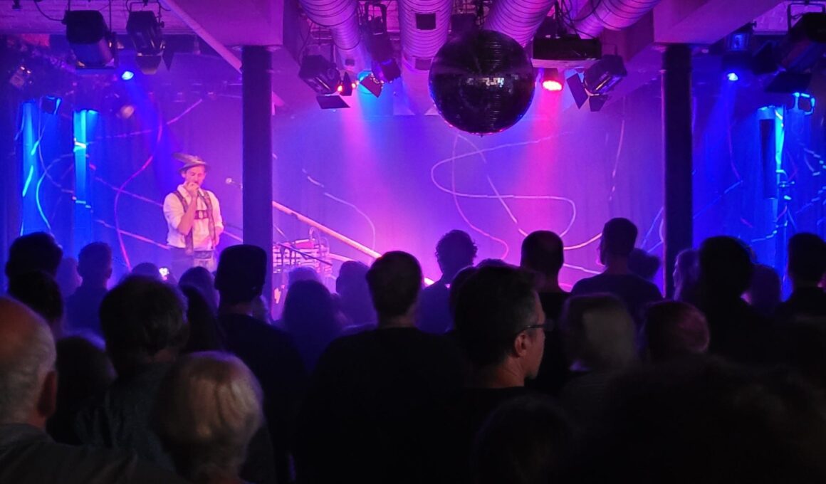 Auf der Bühne des Stuttgarter Kulturzentrums Merlin steht Marcel Engler alias Loisach Marci und musiziert. Er trägt bayerische Tracht. Vor ihm steht ein großes Alphorn. Die Bühne ist bläulich-violett erleuchtet, der Rest des Raums ist dunkel. Im Vordergrund stehen Besucher*innen, manche tanzen. (Foto: vliestext)