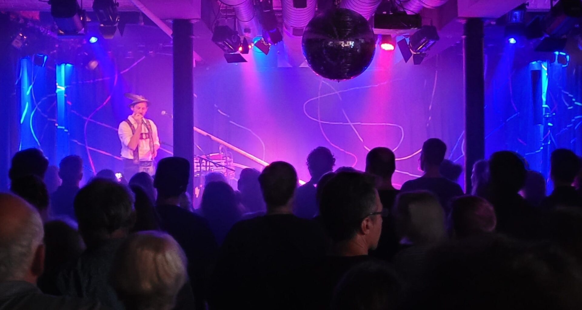 Auf der Bühne des Stuttgarter Kulturzentrums Merlin steht Marcel Engler alias Loisach Marci und musiziert. Er trägt bayerische Tracht. Vor ihm steht ein großes Alphorn. Die Bühne ist bläulich-violett erleuchtet, der Rest des Raums ist dunkel. Im Vordergrund stehen Besucher*innen, manche tanzen. (Foto: vliestext)