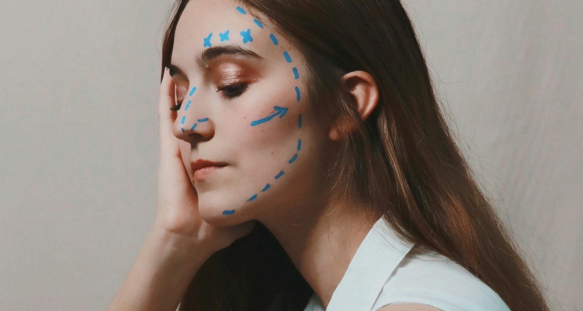 Mensch mit langen braunen Haaren schaut nachdenklich nach unten. Der Kopf ist mit einer Hand abgestützt. Im Gesicht finden sich blaue Markierungslinien, wie man sie von Schönheitsoperationen kennt. (Foto: Joeyy Lee auf Unsplash)