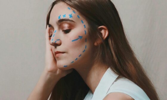 Mensch mit langen braunen Haaren schaut nachdenklich nach unten. Der Kopf ist mit einer Hand abgestützt. Im Gesicht finden sich blaue Markierungslinien, wie man sie von Schönheitsoperationen kennt. (Foto: Joeyy Lee auf Unsplash)