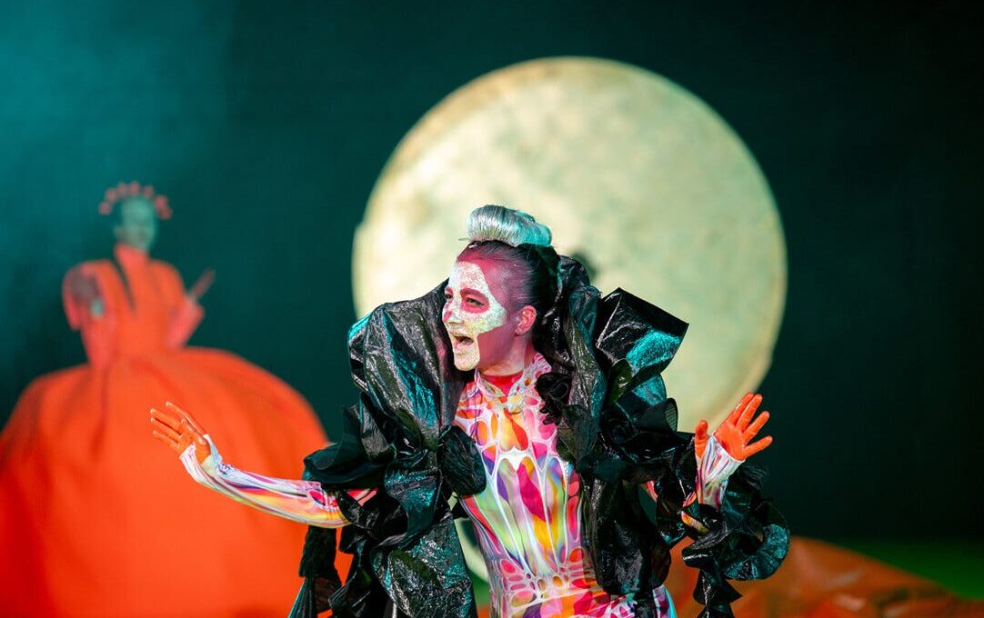 Die Schauspielerin Silvia Schwinger trägt hautenge, bunte Sportkleidung. Um ihre Schultern liegt eine große schwarze Federboa aus Plastik. Ihr ist ein Totenkopf ins Gesicht geschminkt. Sie hat die rot gefärbten Hände erhoben und blickt sprechend nach rechts. (Foto: Björn Klein)