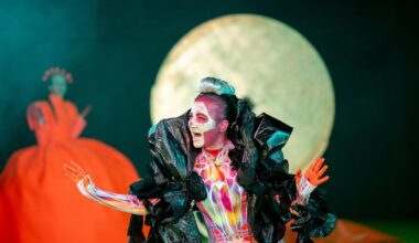 Die Schauspielerin Silvia Schwinger trägt hautenge, bunte Sportkleidung. Um ihre Schultern liegt eine große schwarze Federboa aus Plastik. Ihr ist ein Totenkopf ins Gesicht geschminkt. Sie hat die rot gefärbten Hände erhoben und blickt sprechend nach rechts. (Foto: Björn Klein)