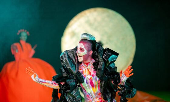 Die Schauspielerin Silvia Schwinger trägt hautenge, bunte Sportkleidung. Um ihre Schultern liegt eine große schwarze Federboa aus Plastik. Ihr ist ein Totenkopf ins Gesicht geschminkt. Sie hat die rot gefärbten Hände erhoben und blickt sprechend nach rechts. (Foto: Björn Klein)