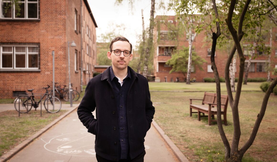 Titelbild: In dunkler Kleidung steht Paul Bokowski, der Autor von „Schlesenburg“, vor Häusern im Innern der Berliner Schillerpark-Siedlung und schaut in die Kamera.