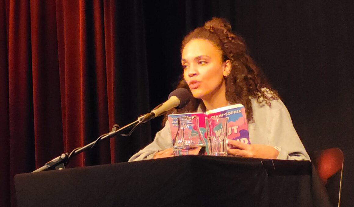 Ciani-Sophia Hoeder sitzt vor dunklem Hintergrund an einem schwarzen Tisch mit Mikrofon. In Händen hält sie ihr Buch „Wut und Böse“.