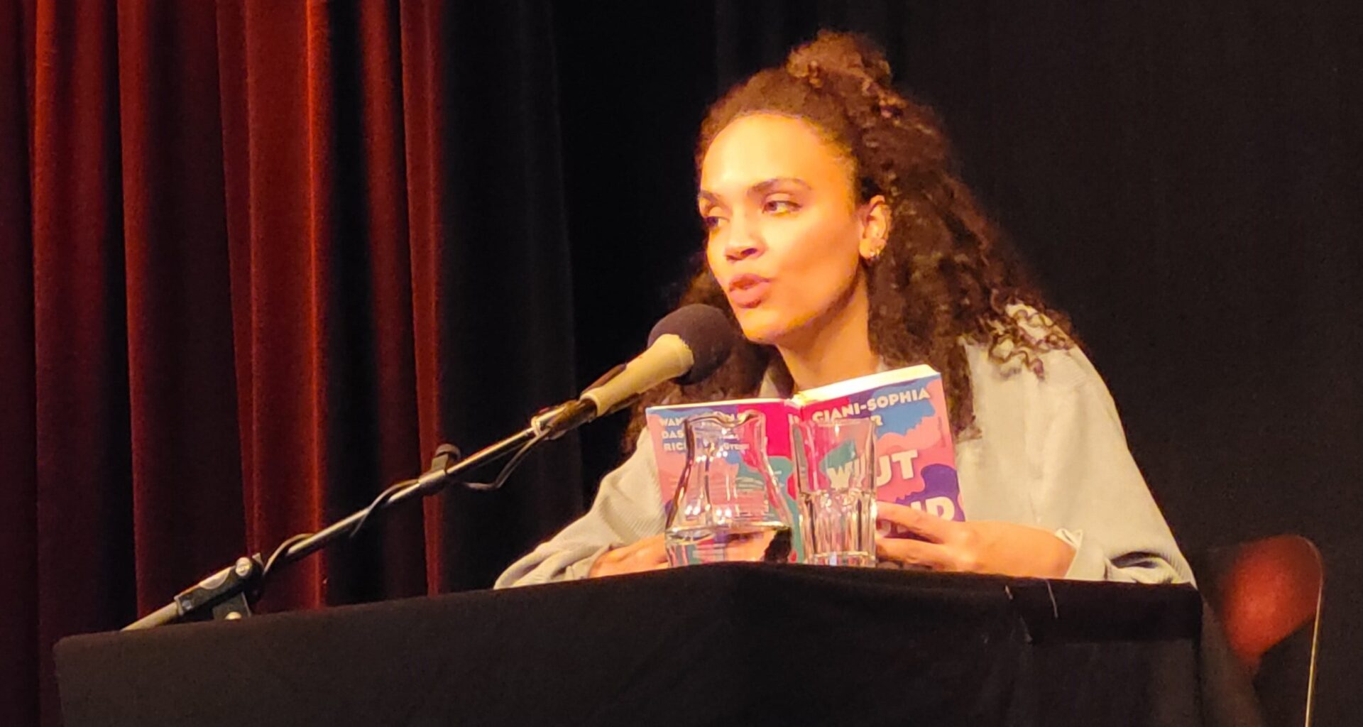 Ciani-Sophia Hoeder sitzt vor dunklem Hintergrund an einem schwarzen Tisch mit Mikrofon. In Händen hält sie ihr Buch „Wut und Böse“.