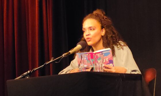 Ciani-Sophia Hoeder sitzt vor dunklem Hintergrund an einem schwarzen Tisch mit Mikrofon. In Händen hält sie ihr Buch „Wut und Böse“.