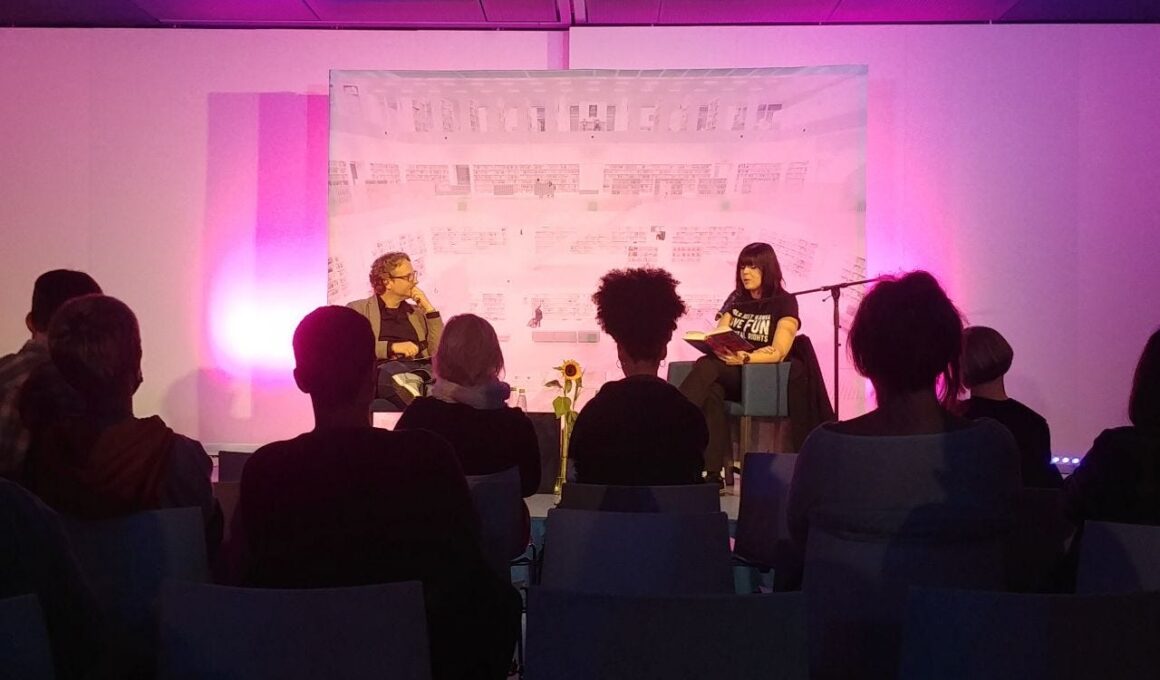 Lesung mit zwei Menschen auf in rosa Neon-Licht erleuchteter Bühne. Im Vordergrund sitzen Zuschauer*innen, deren Rücken zu sehen ist. Auf der Bühne liest die Autorin Mareike Fallwickl aus ihrem Roman "Die Wut, die bleibt". Der Moderator Frank Rudkoffsky hört zu. (Foto: vliestext)